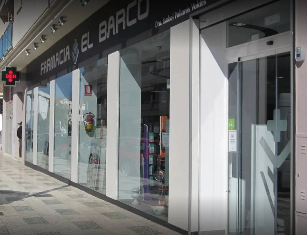 Fachada acristalada de la Farmacia El Barco con su rótulo y la cruz verde encendida