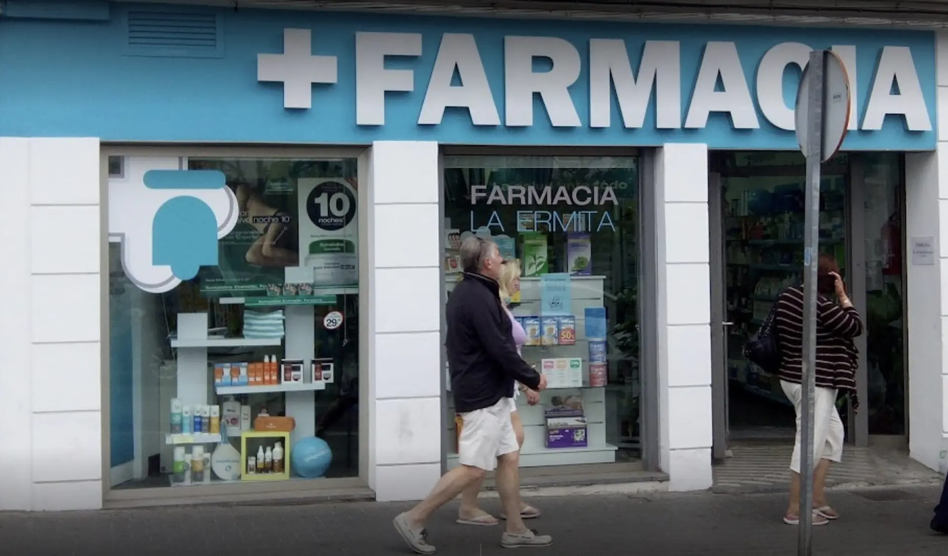 Fachada de la farmacia con la cruz y el rótulo azul, y gente paseando por la acera