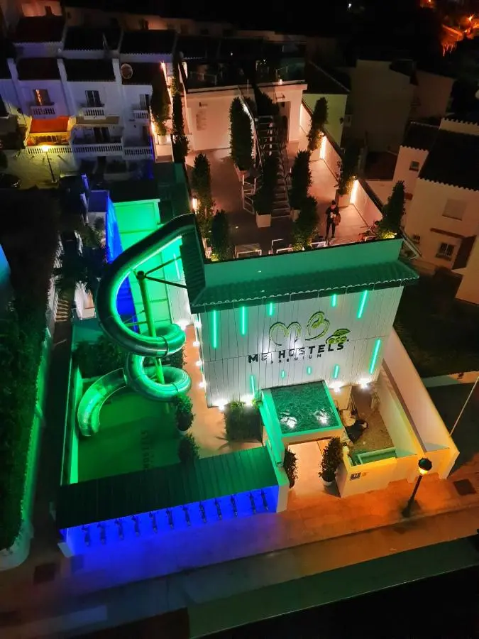 Vista cenital nocturna del edificio con el tobogán iluminado y la azotea con cipreses