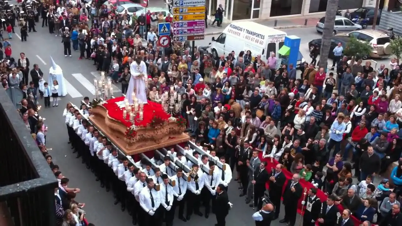 Portadores de blanco con un trono de alfombra roja avanzando entre el público, vistos desde un balcón