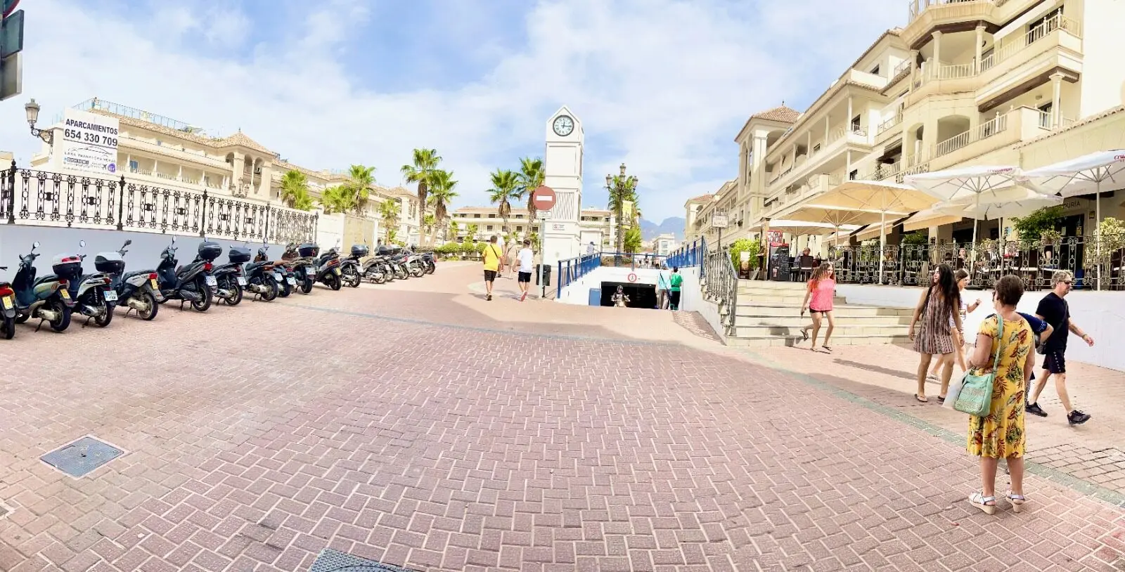 Motos aparcadas, reloj y escaleras de acceso con gente paseando junto a las terrazas