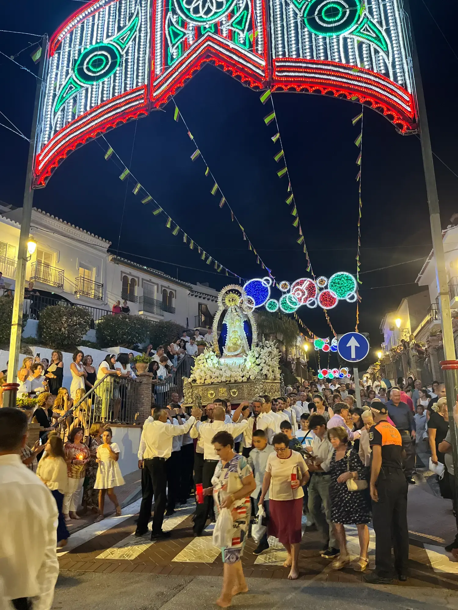 Procesión de la Virgen bajo un arco de luces de feria, con vecinos llenando la calle