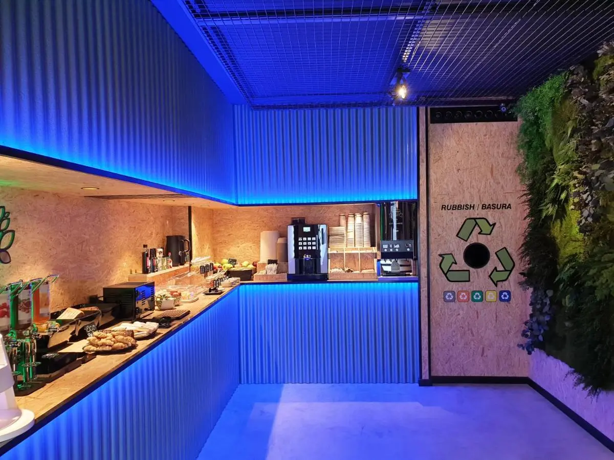 Zona de desayuno con cafetera, bufé y luces azules, junto a un punto de reciclaje