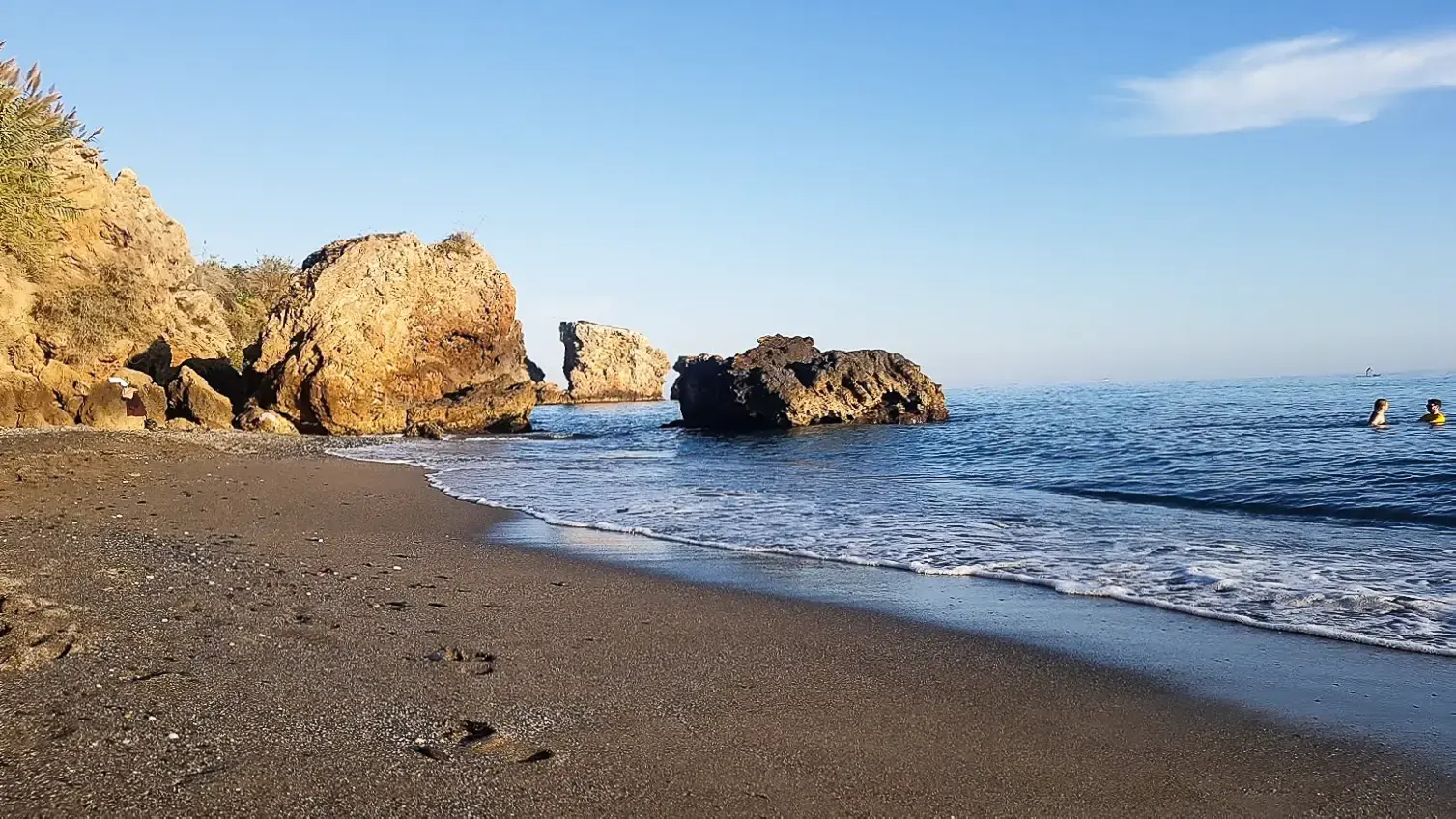 Orilla de arena oscura y grandes rocas en La Caleta de Maro, con bañistas al atardecer