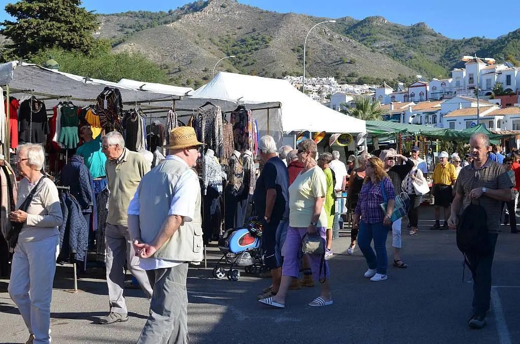 Visitantes paseando entre puestos de ropa en el mercadillo de los martes, con la sierra detrás