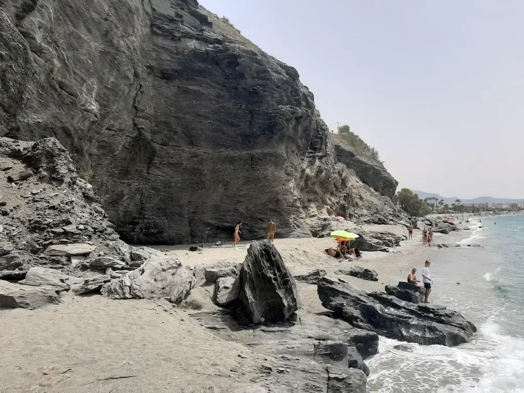Rocas oscuras y acantilado al final de la playa El Playazo, con bañistas