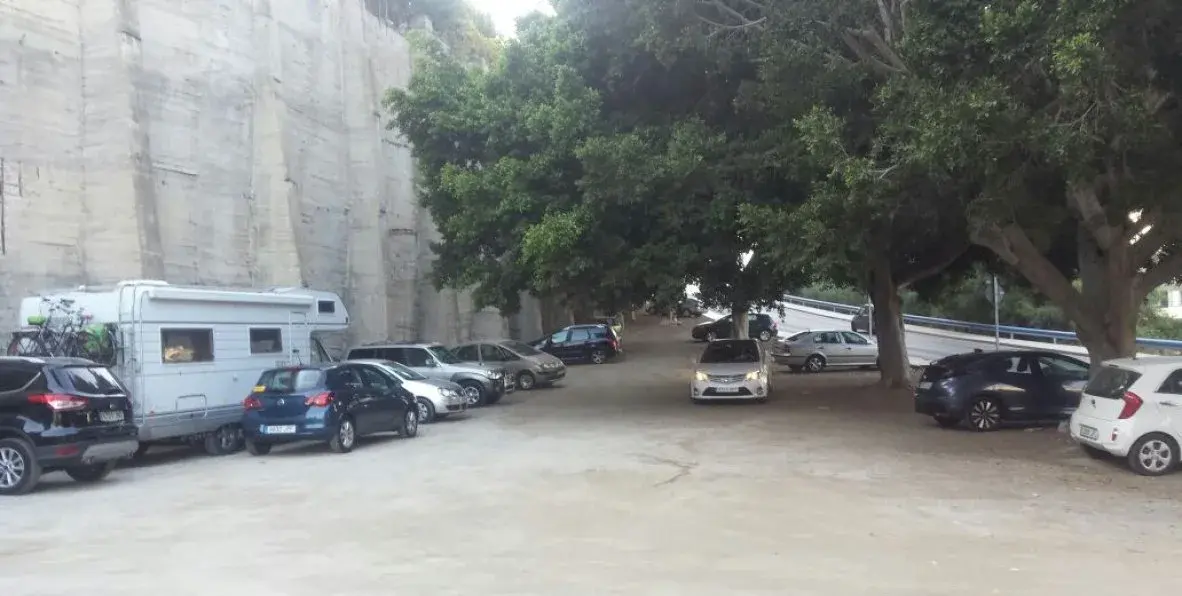 Aparcamiento de tierra junto al muro de hormigón, con autocaravana y coches bajo los árboles