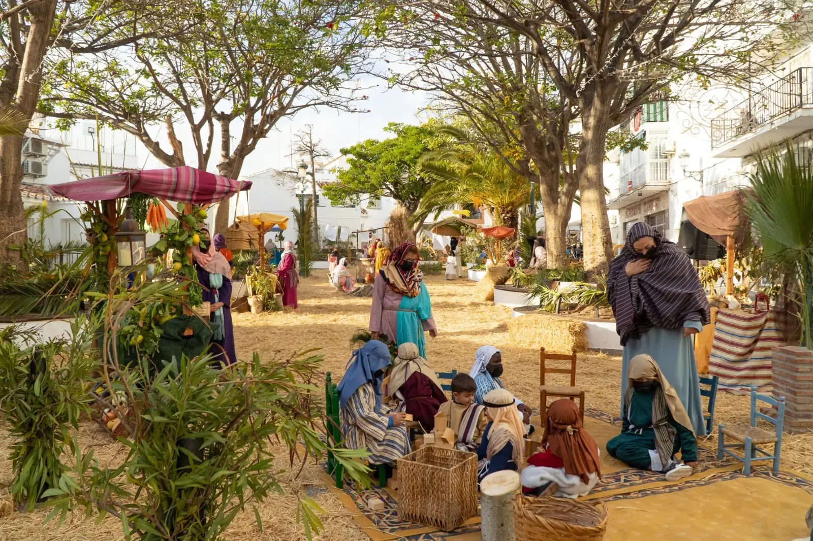 Escena de aldea con figurantes vestidos de época sobre paja bajo los árboles de la plaza