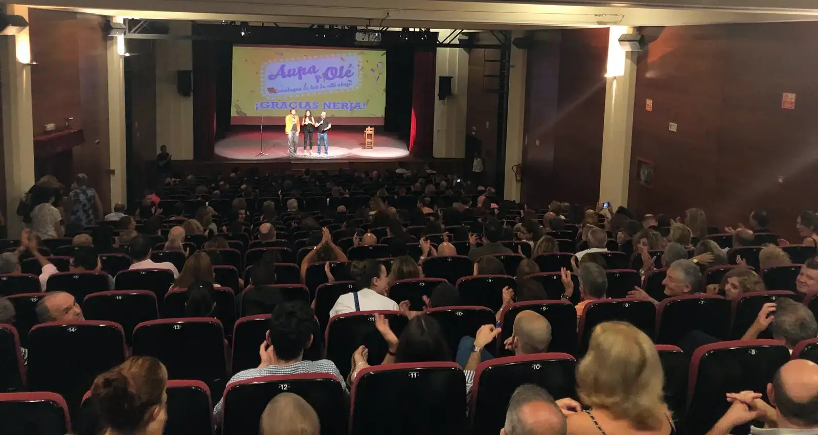 Público aplaudiendo en el auditorio lleno, con tres personas en el escenario