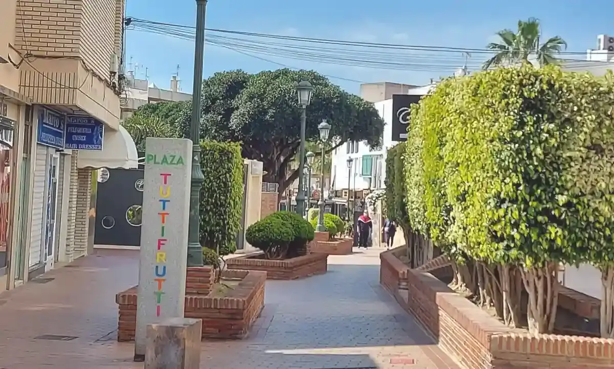 Pilar con el nombre de la plaza en letras de colores a la entrada, entre setos y jardineras