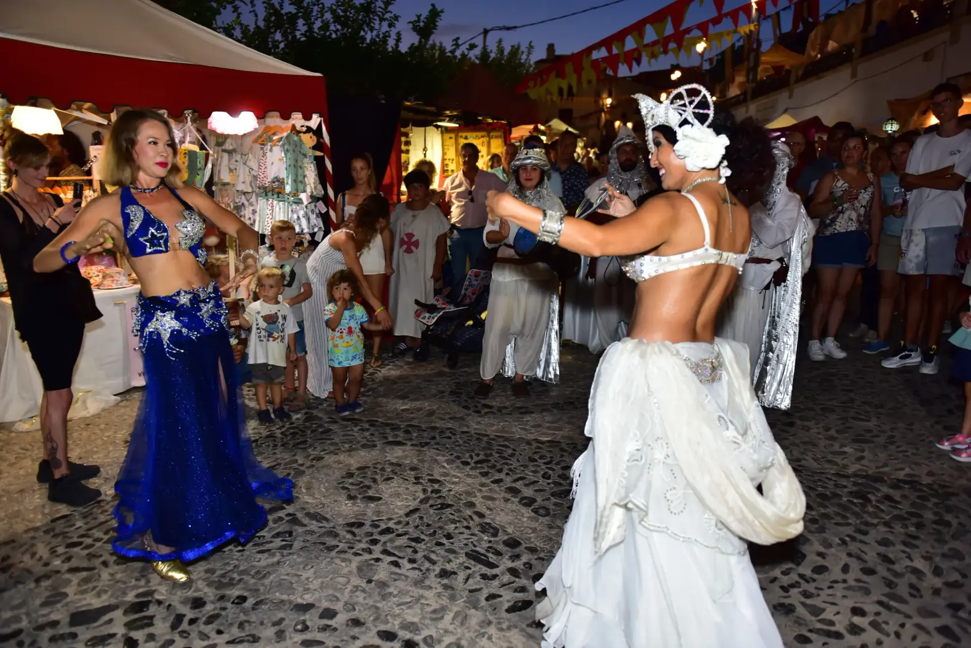 Dos bailarinas de danza oriental actuando de noche entre puestos y público del festival