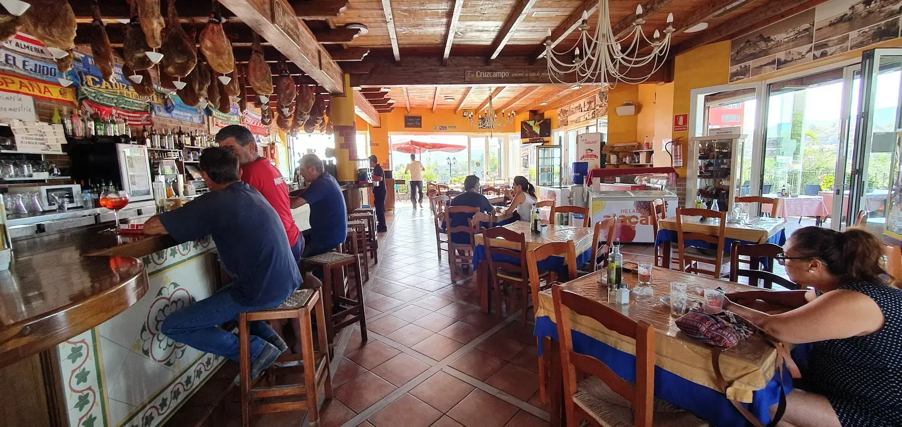 Bar restaurante con barra, jamones colgados del techo y mesas con manteles junto a los ventanales