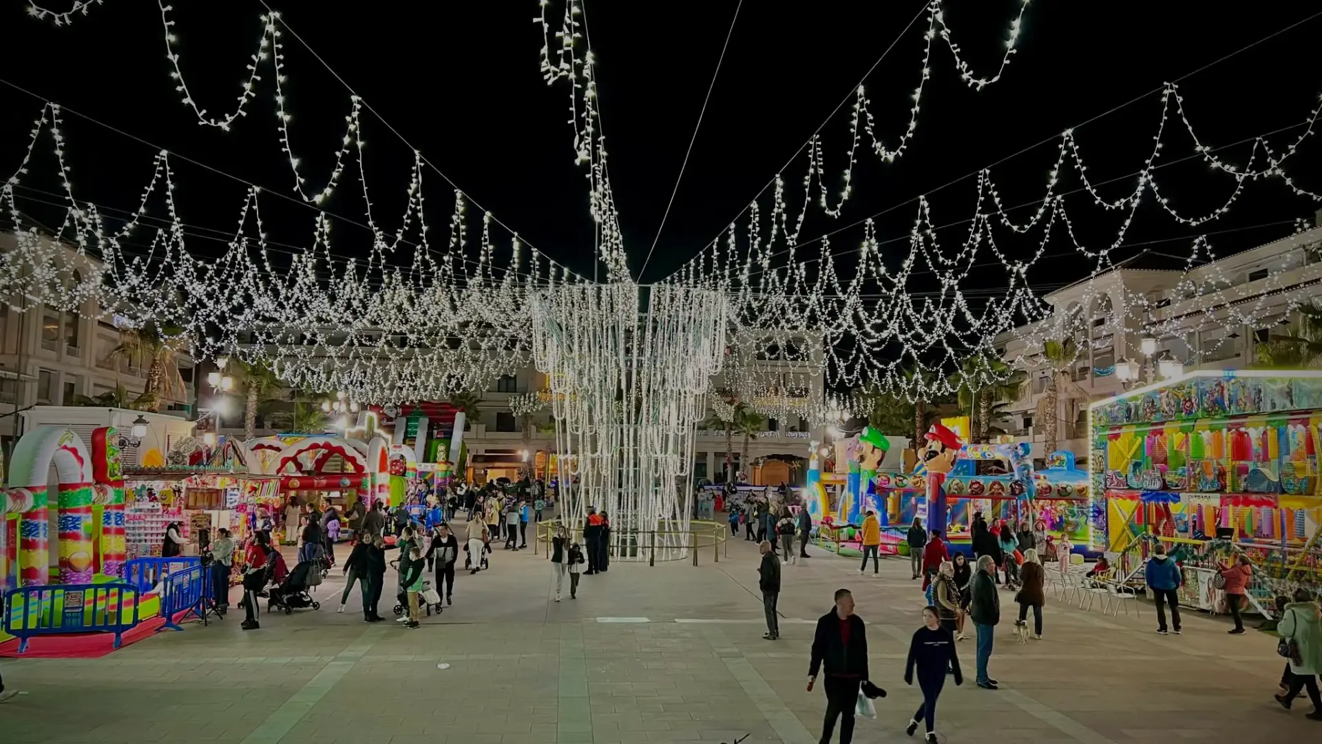 Plaza con guirnaldas de luces navideñas, atracciones infantiles y gente paseando de noche