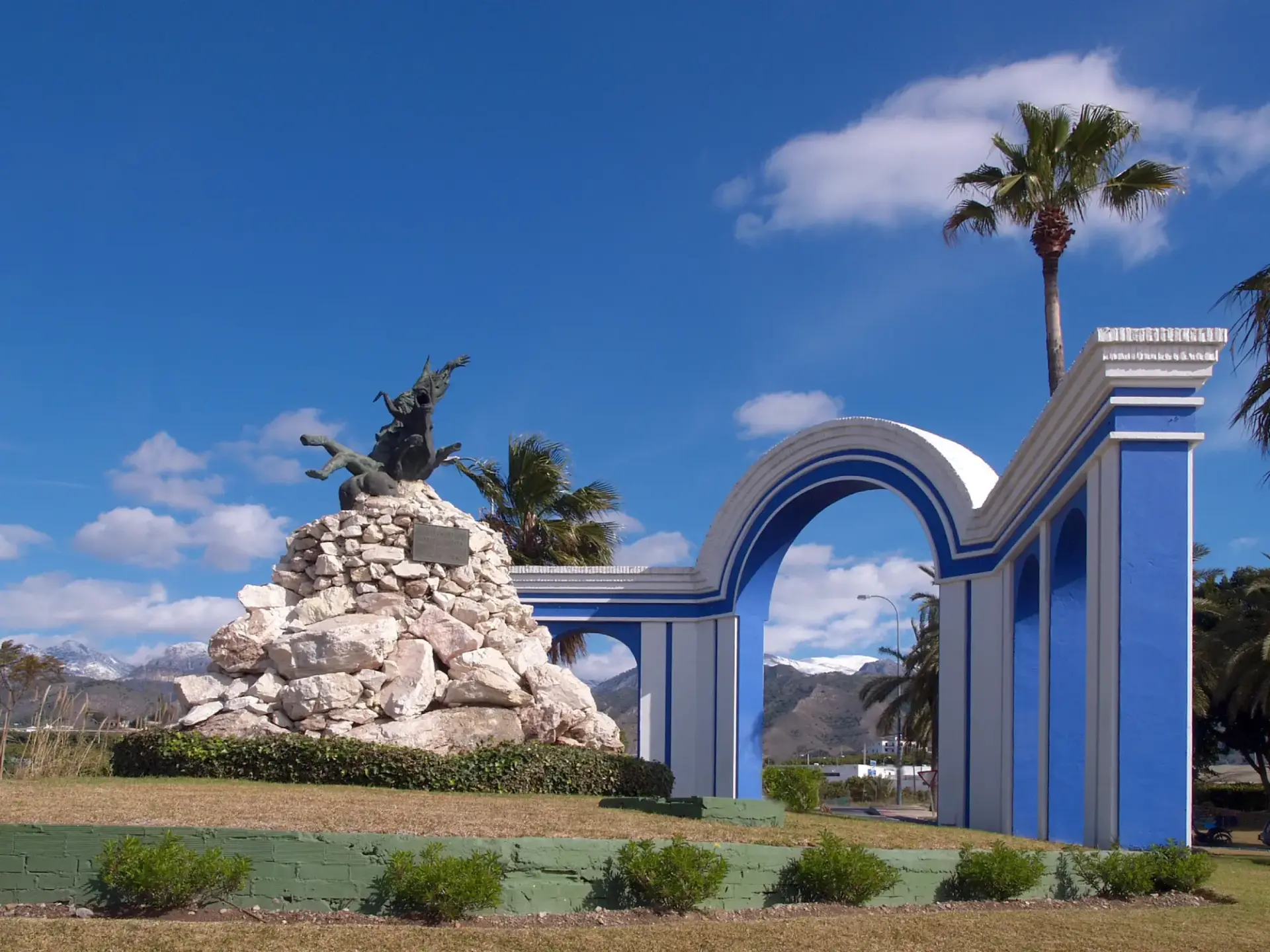 Escultura sobre un montículo de piedras junto a los arcos azules, con sierras nevadas al fondo