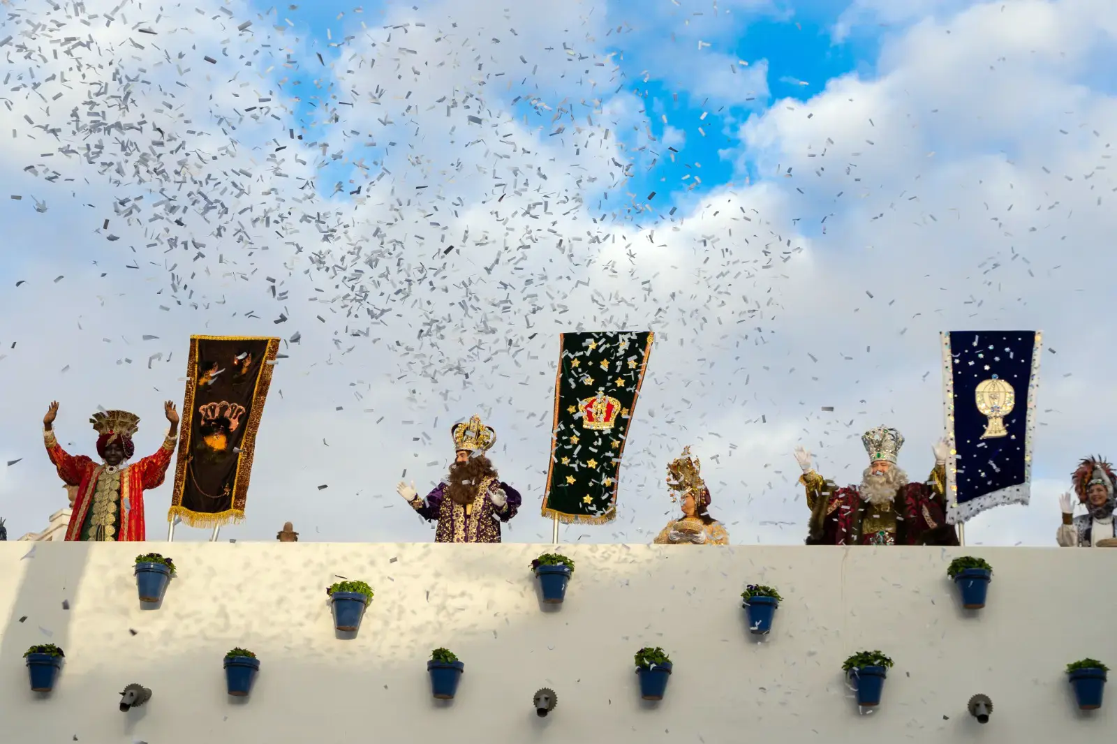 Los Reyes Magos saludan desde lo alto de un muro blanco entre estandartes y lluvia de confeti