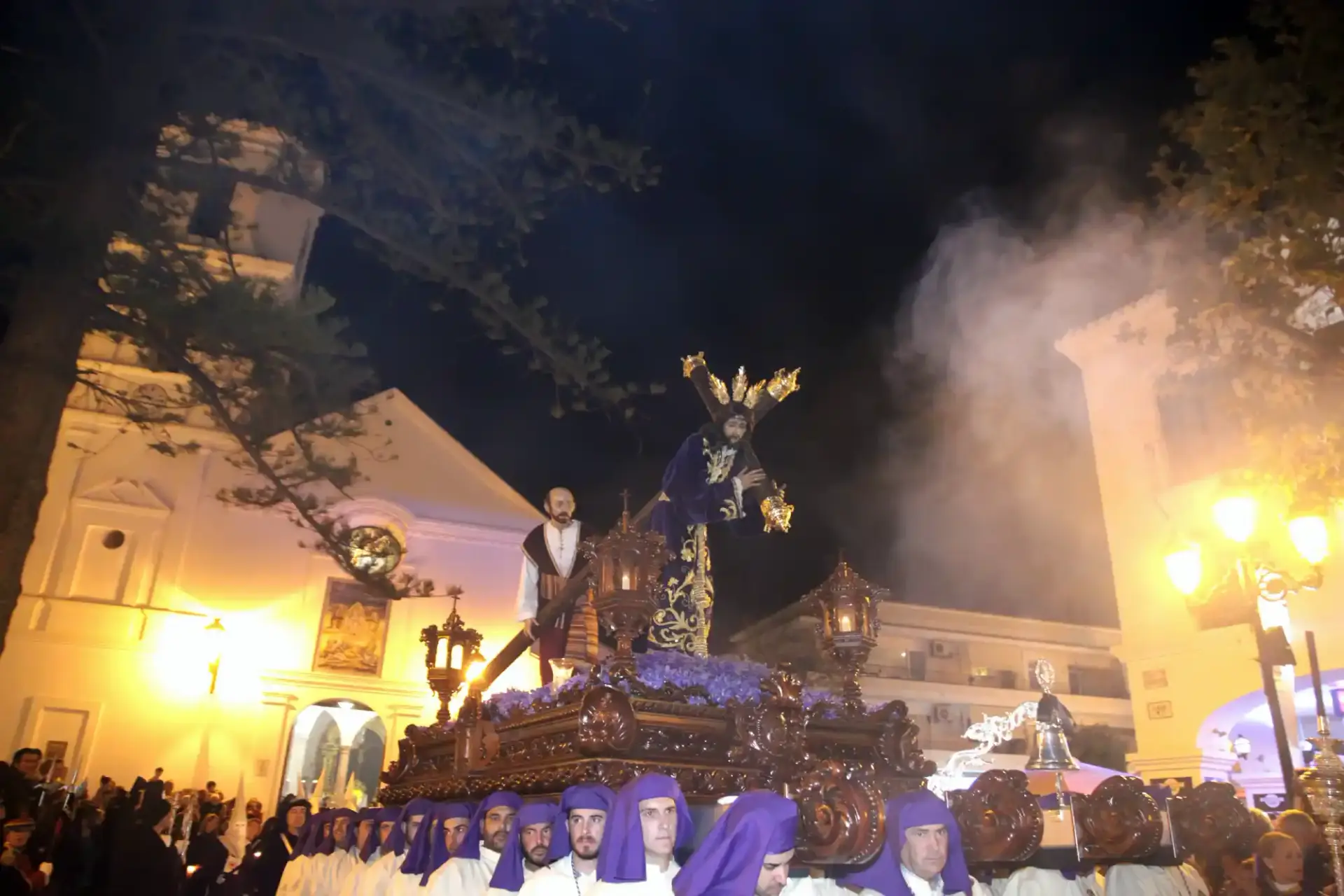 Imagen de Jesús con la cruz y túnica morada en procesión nocturna, con portadores de capirote morado