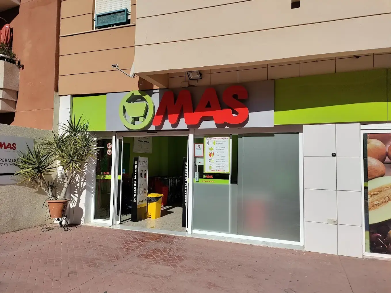 Entrada del supermercado con el logo del carrito, puerta automática abierta y una palmera en maceta