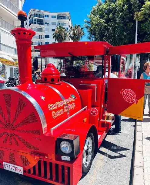Locomotora roja del tren turístico con el rótulo City Sightseeing Cueva de Nerja, parada en la calle