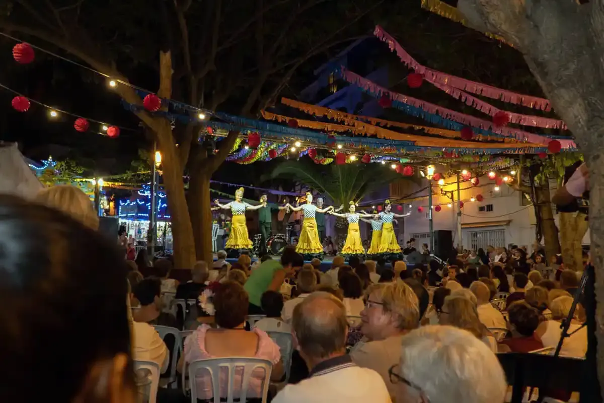 Bailarinas con vestidos amarillos en el escenario ante el público sentado, de noche y con farolillos
