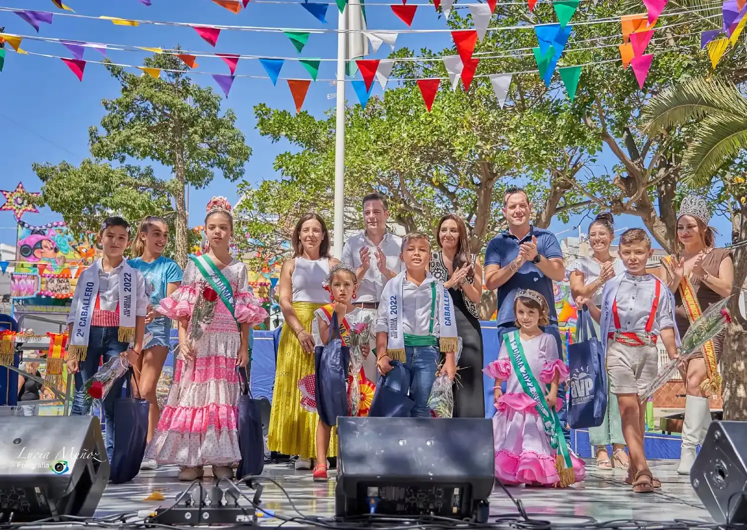 Niños con bandas y trajes de flamenca en el escenario junto a autoridades, bajo banderines de colores