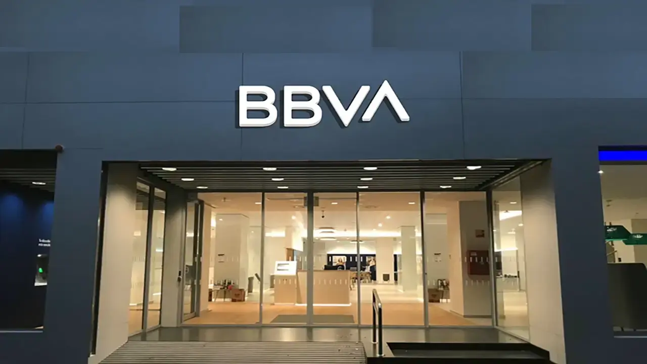 Entrada acristalada de una oficina BBVA con el logotipo iluminado sobre fachada gris