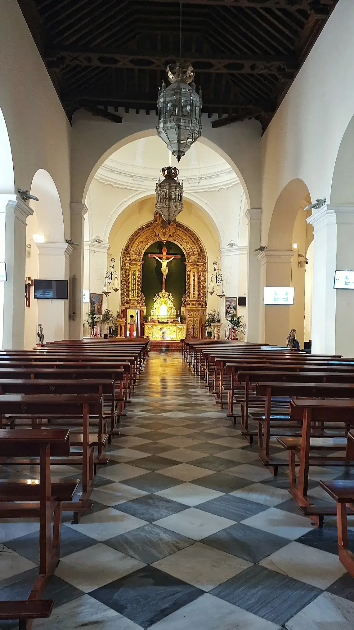 Nave central con bancos de madera, suelo ajedrezado y retablo dorado con crucifijo al fondo
