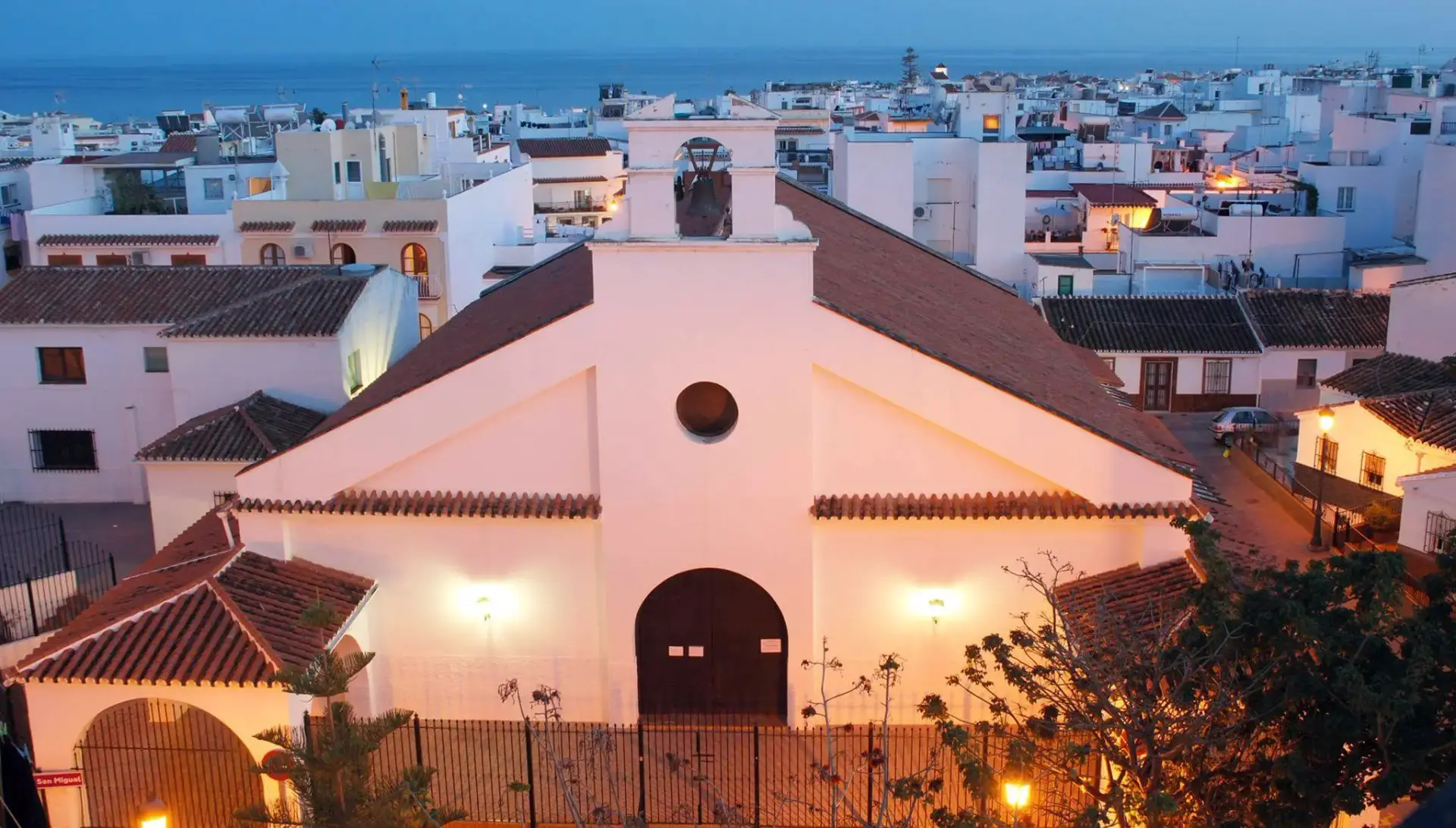 Fachada iluminada de la Iglesia San Miguel al anochecer, con los tejados de Nerja y el mar