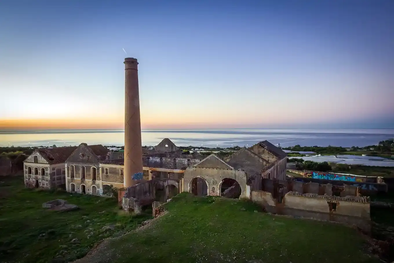 Ruinas de la fábrica y su alta chimenea al atardecer, con el mar en el horizonte