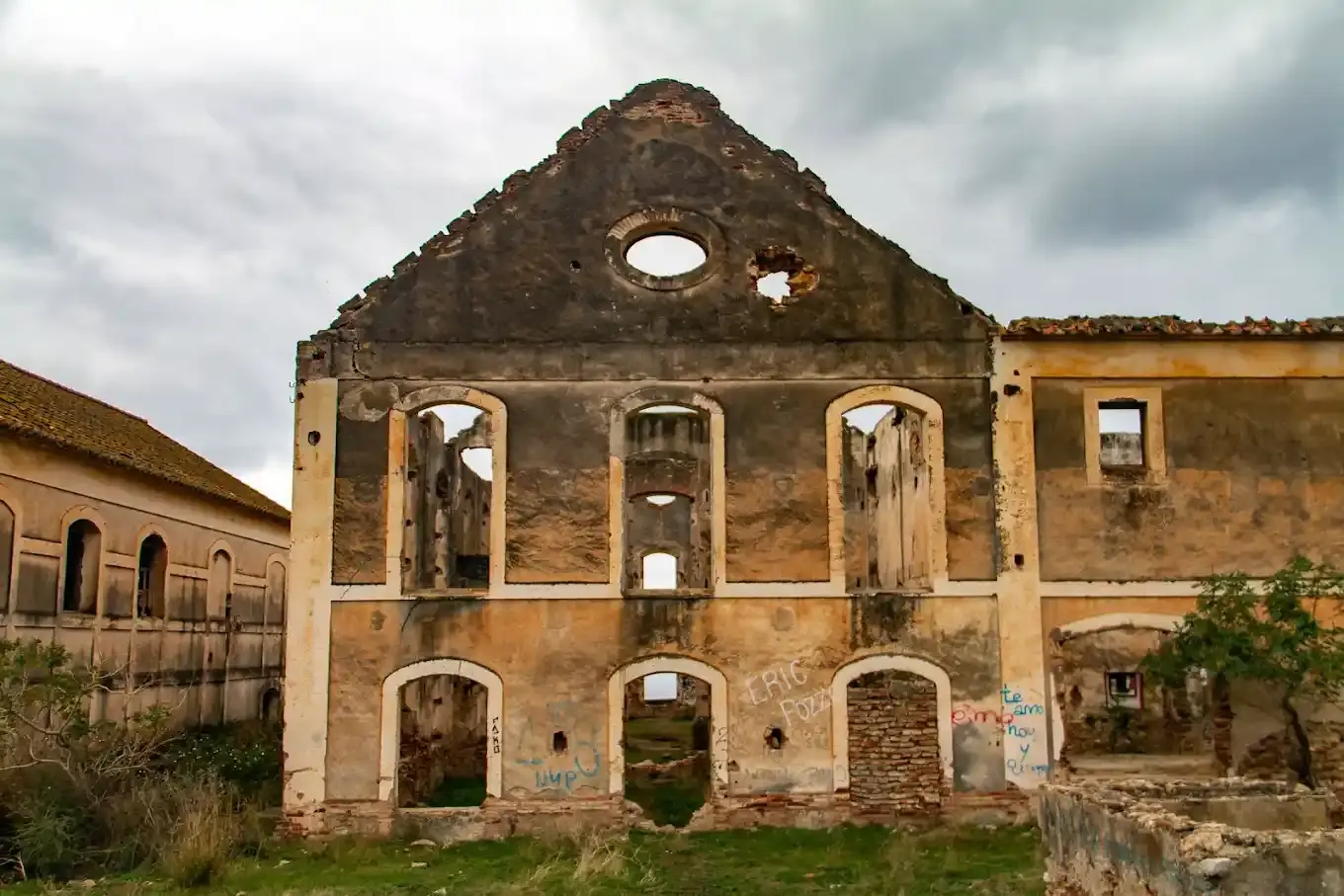 Fachada vacía de una nave con óculo y ventanas sin cristales bajo un cielo nublado