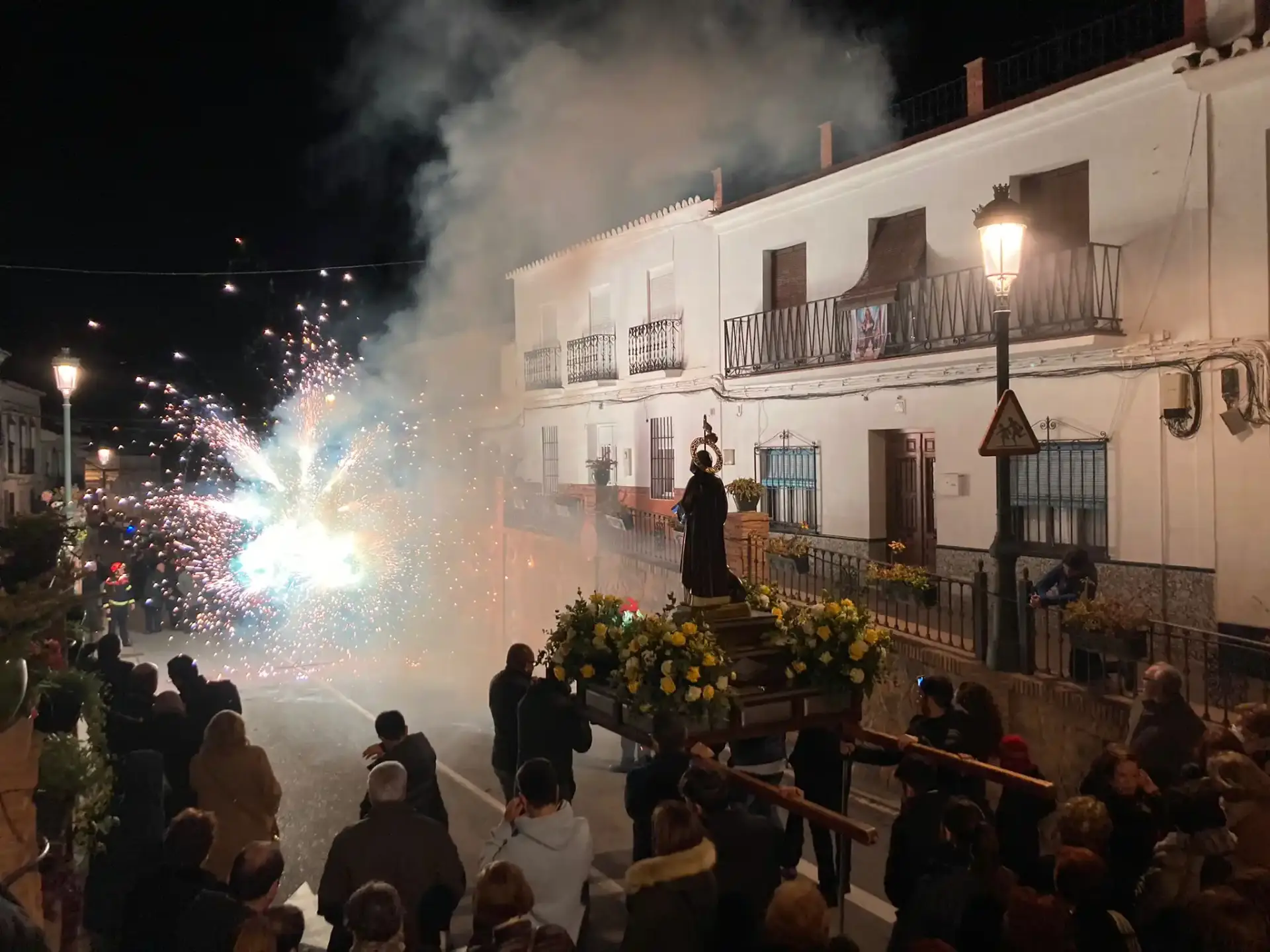 Procesión de San Antón entre fuegos artificiales y humo, con el trono adornado de flores amarillas