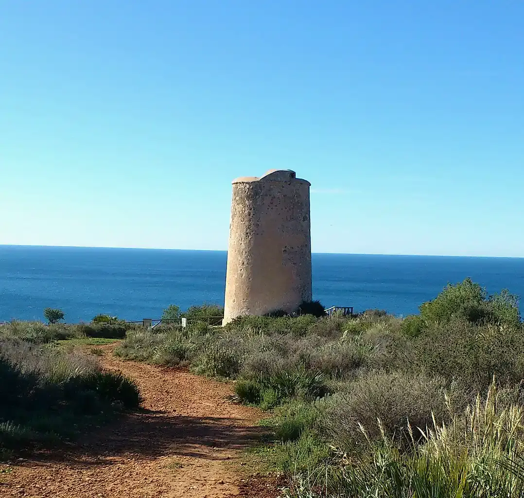 Camino de tierra entre matorral que lleva a la torre vigía, con el mar azul detrás