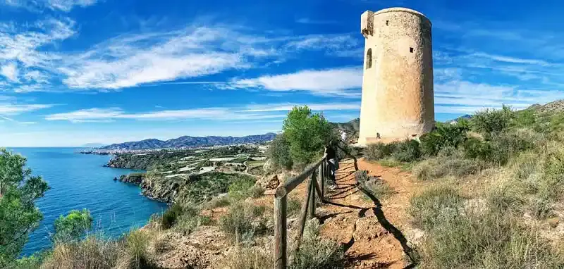 Sendero con barandilla de madera junto a la torre, con la costa y las sierras al fondo