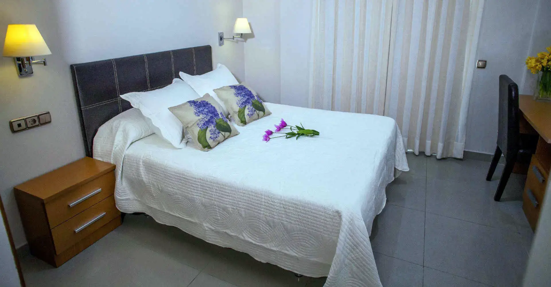 Cama de matrimonio con cabecero gris, cojines de flores lilas y mesilla de madera