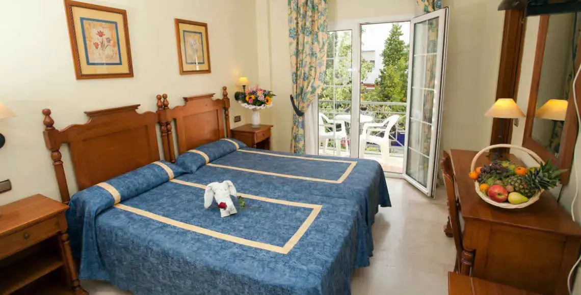 Habitación con dos camas de madera, colchas azules, frutero y terraza con sillas blancas