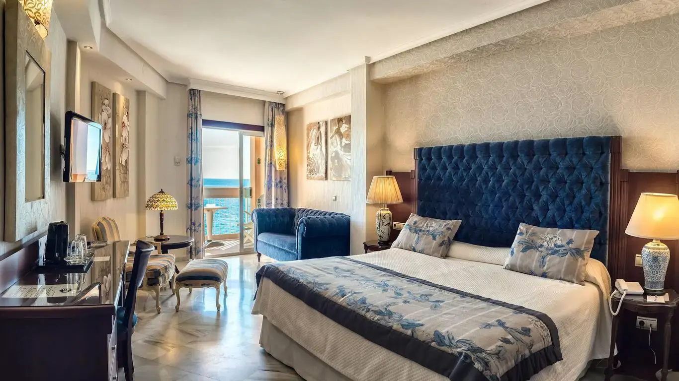 Habitación con cabecero acolchado azul, sillón, escritorio y terraza abierta al mar