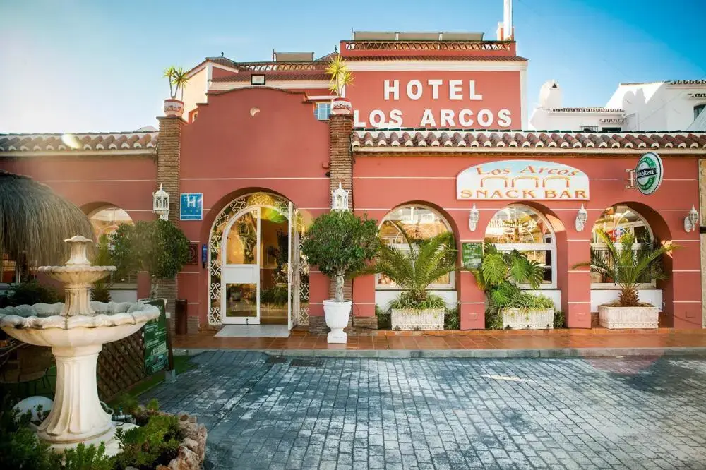 Fachada roja con arcos, rótulo de Los Arcos Snack Bar y una fuente en la entrada
