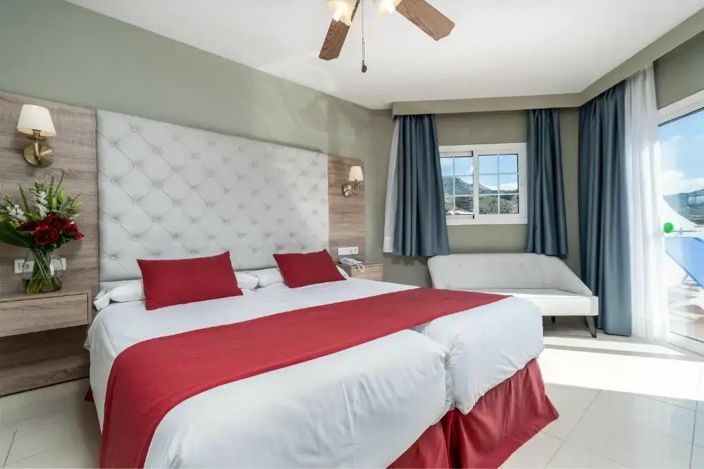 Habitación con camas juntas, cabecero capitoné blanco, colcha roja y ventilador de techo