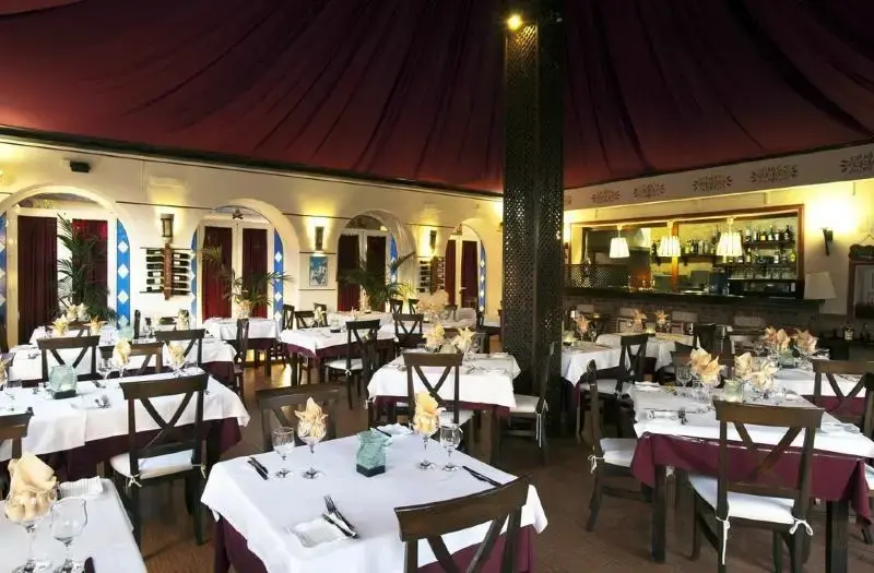Restaurante con mesas de mantel blanco, servilletas dobladas, techo de tela granate y barra