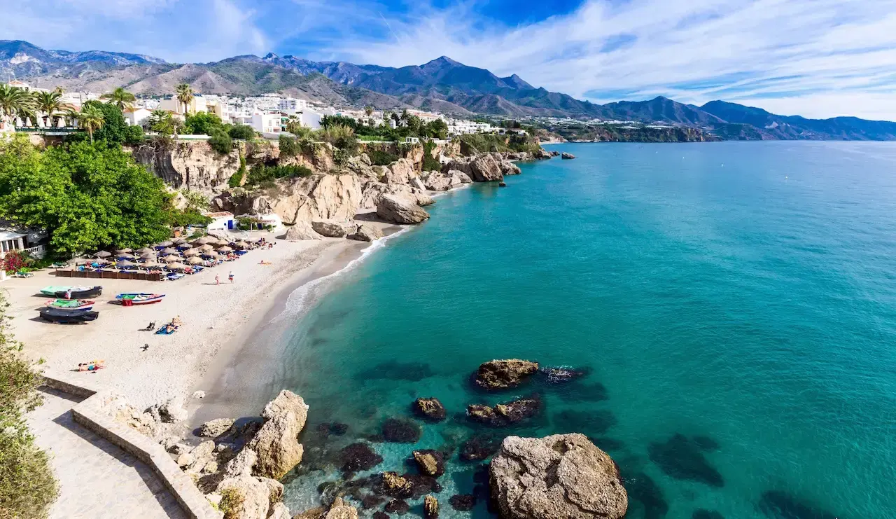 Cala de arena con barcas, sombrillas y rocas en el agua turquesa, con la sierra detrás