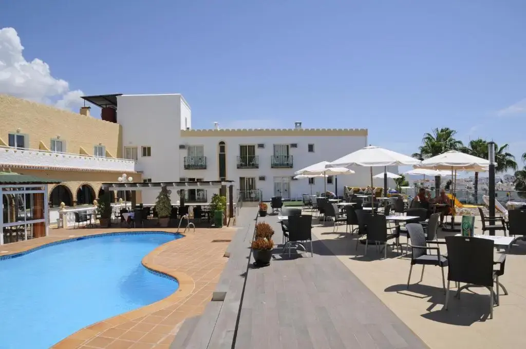 Terraza con mesas y sombrillas junto a la piscina y el edificio blanco del hotel