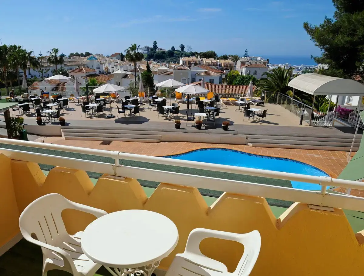Balcón con mesa y sillas blancas sobre la piscina y la terraza, con el mar al fondo