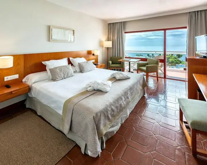 Habitación con cama de matrimonio, albornoces, sillones y puerta a una terraza con vistas al mar