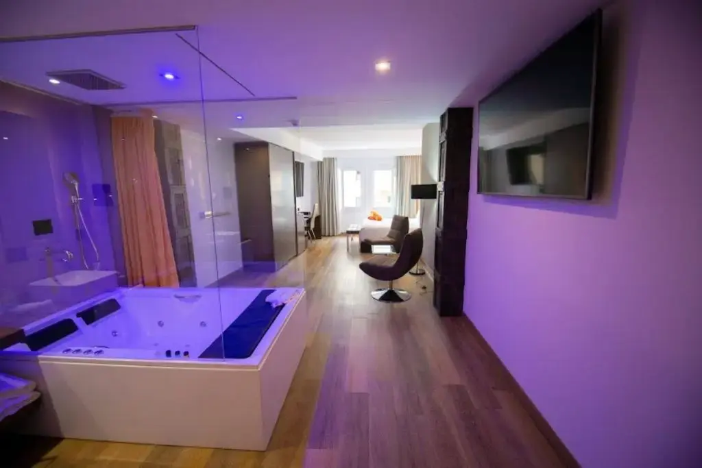 Suite con bañera de hidromasaje tras un cristal, luz morada, televisor y sillón