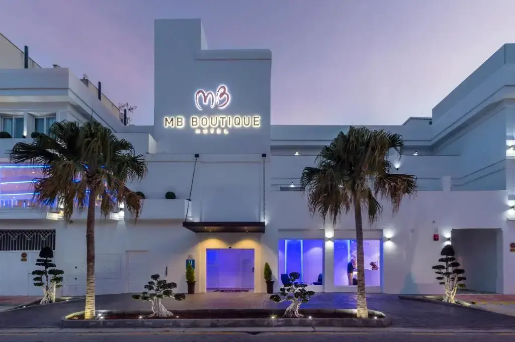 Fachada blanca del MB Boutique Hotel al anochecer, con letrero iluminado y palmeras