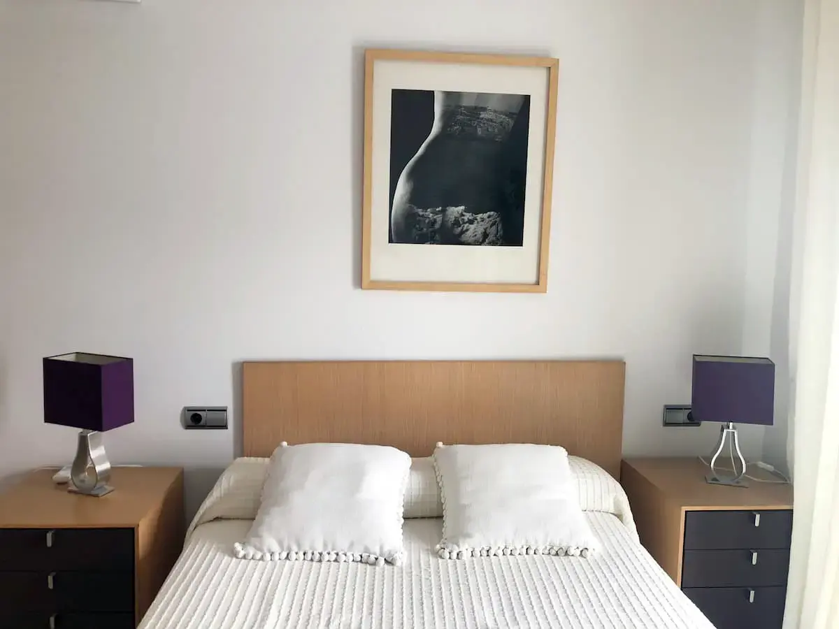 Dormitorio con cama de matrimonio, cabecero de madera, mesillas con lámparas moradas y cuadro