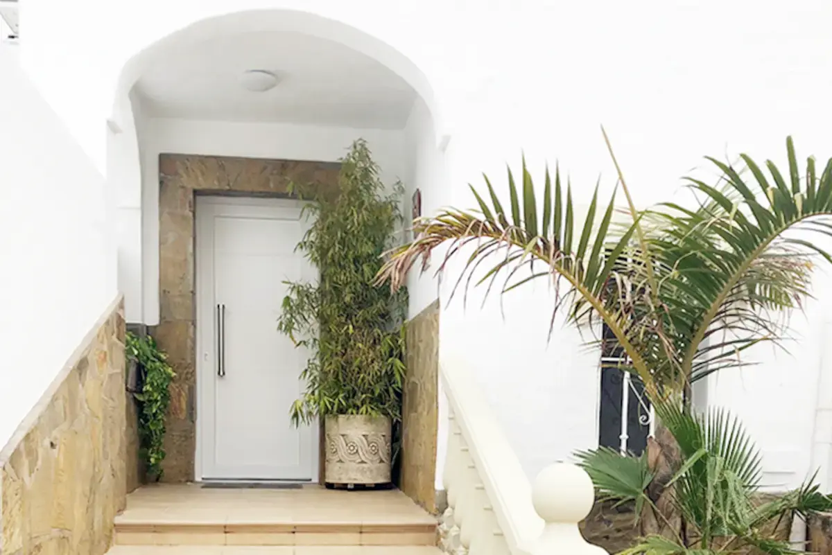 Entrada al Apartamento Condal con puerta blanca bajo un arco, plantas y una palmera
