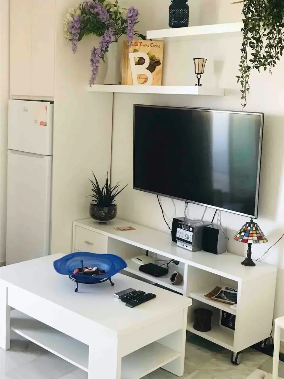 Rincón del salón con televisor de pantalla plana, mueble blanco, estantes y frigorífico al lado