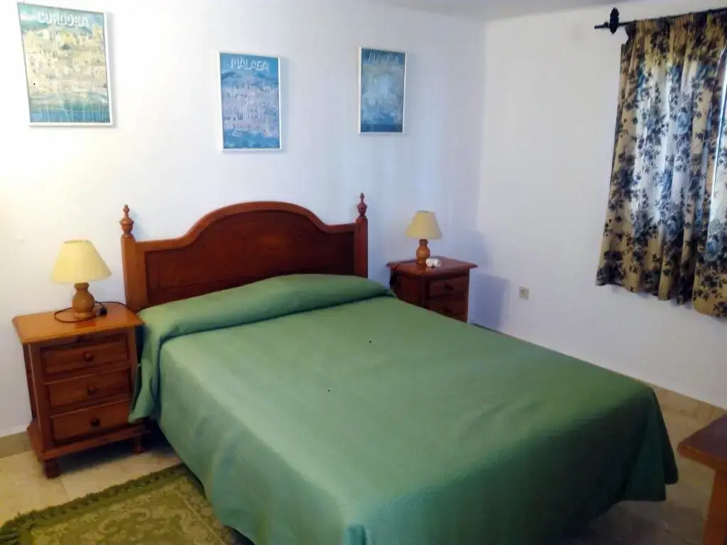 Dormitorio con cama de matrimonio, colcha verde, mesitas de madera y láminas en la pared