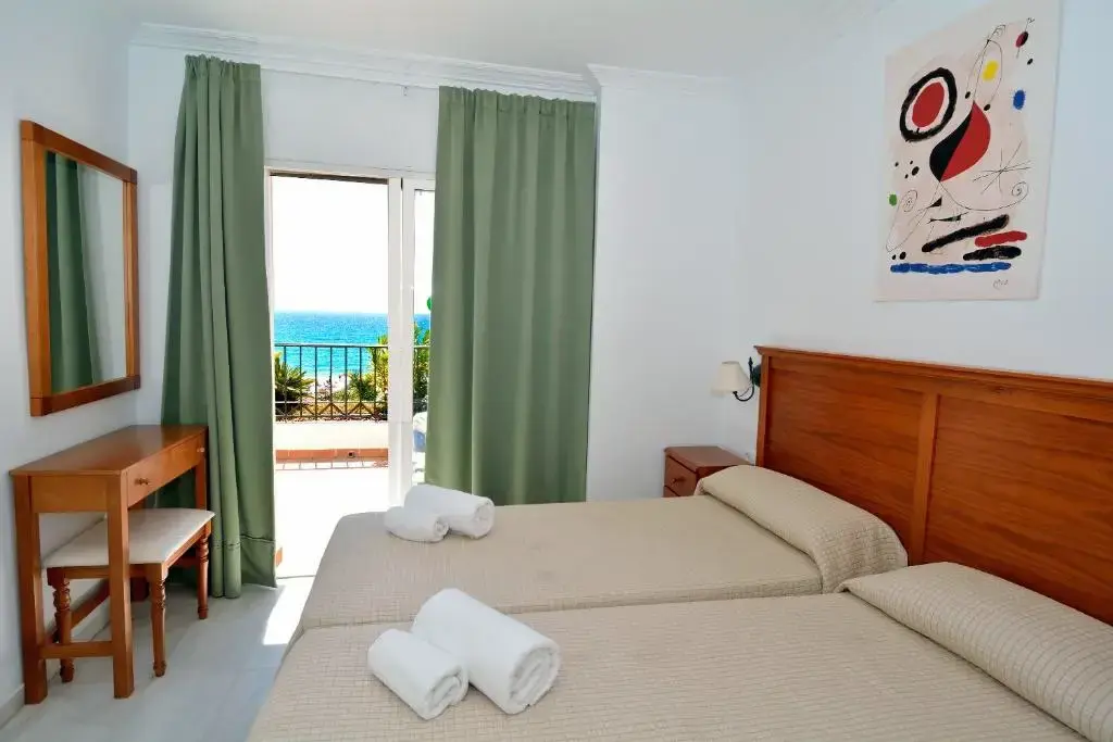 Habitación con dos camas, toallas, tocador con espejo y balcón abierto al mar