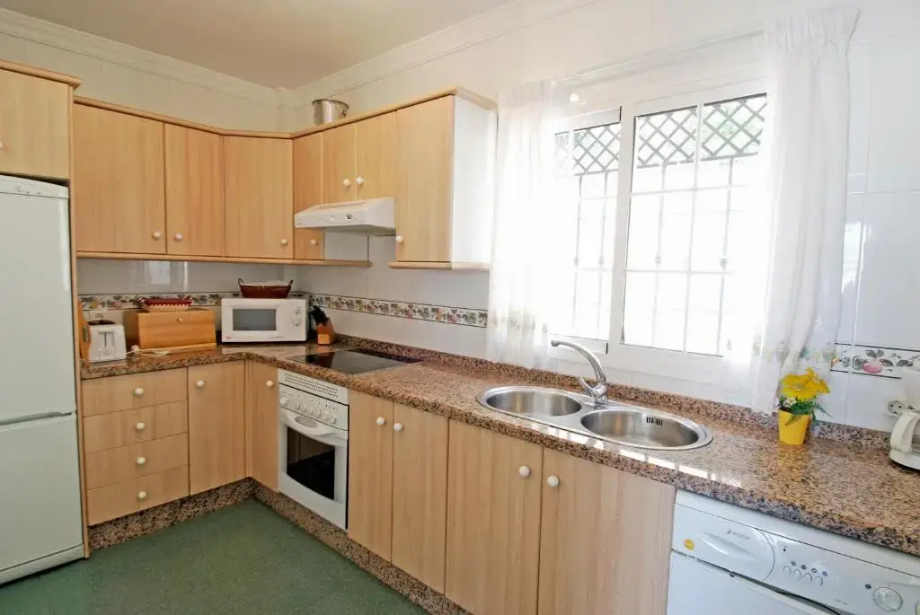 Cocina con muebles de madera clara, fregadero doble, horno, microondas y ventana con reja